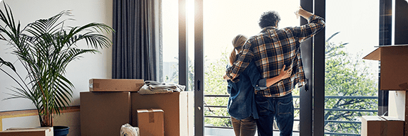 Photo d’un homme et d’une femme enlacés devant la fenêtre de leur nouveau logement avec des boîtes de déménagement 