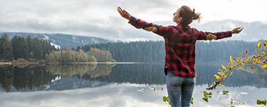 Jeune femme ouvre grand les bras devant un lac entouré d’une forêt