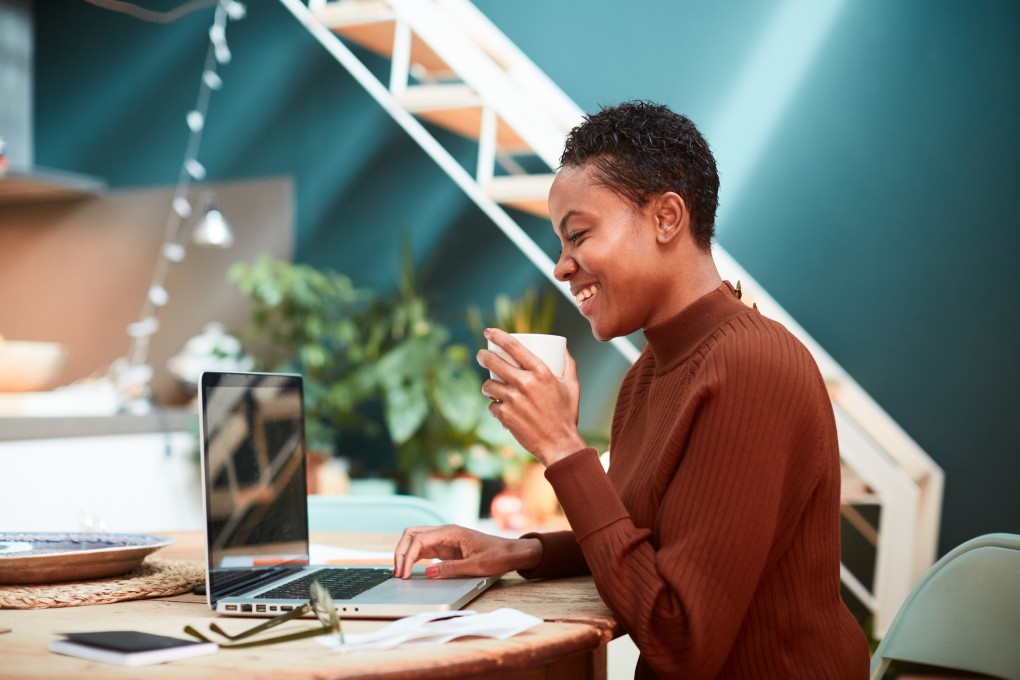 Une femme en télétravail prend son café en souriant devant son ordinateur.